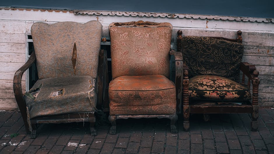 A row of three antique armchairs and a vintage wooden armchair with fabric upholstery and ornate carved legs are positioned outside against a weathered brick wall and wooden cladding. The leftmost chair features a beige, floral-patterned fabric that is torn and frayed, revealing the inner stuffing and wooden frame beneath. The middle chair has an orange, patterned fabric covering, showing signs of wear with some fading and minor tears. The rightmost piece is a wooden armchair with a dark, intricate fabric cushion on the seat and backrest, supported by turned wooden spindles on the armrests and legs. The chairs are situated on a brick pavement, and the background includes a slightly peeling, white-painted wooden panel. This setting suggests the furniture is awaiting removal or disposal, relevant to home relocation or furniture transport services offered by Man With a Van Pimlico, which specialises in removals and moving logistics.