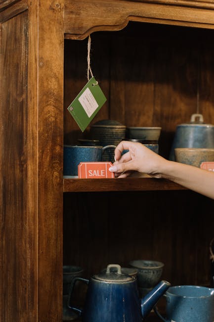 A person's hand is shown removing a small orange sale tag from a wooden shelf in an interior setting, which contains a collection of ceramic canisters and jugs in shades of blue, green, and black. The shelf is part of a wooden cabinet or display unit, with the interior lit by natural or warm lighting. Hanging above the shelf is a string with a green price tag attached. The scene suggests a shop or storage area, possibly related to packing or preparing household items for a move. The image is associated with house removals and furniture transport, reflecting the process of packing and organizing belongings during a home relocation, consistent with the services offered by Man With a Van Pimlico.