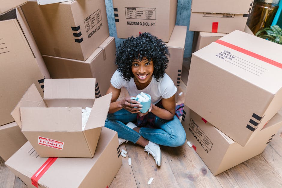 A woman with curly black hair and a bright smile is seated on a wooden floor surrounded by cardboard boxes used for home packing and moving. She is holding a blue mug filled with a beverage in her hands. The boxes vary in size and are sealed with packing tape, some with labels indicating contents or fragile warnings. One open box in front of her contains crumpled packing paper, while others are stacked around her, some upright and others lying on their sides. The scene appears to be indoors, in a room with natural light, and the boxes are likely prepared for a house relocation or moving process. The presence of packing materials, boxes, and the woman's engaged expression reflect the active packing phase of a professional or personal move, aligning with services offered by Man With a Van Pimlico for efficient furniture transport and home removals.