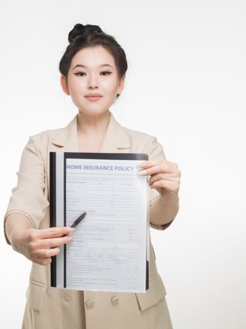 A woman with dark hair styled in an updo, dressed in a beige blazer, stands against a plain white background, holding a clipboard with a visible document titled 'Home Insurance Policy'. She points to the clipboard with a pen in her right hand, indicating a focus on insurance coverage related to house moves. The scene emphasizes the importance of understanding insurance and liability during home relocation or furniture transport, aligning with typical house removal services. The professional appearance and neutral setting support the theme of organised, secure moving logistics.
