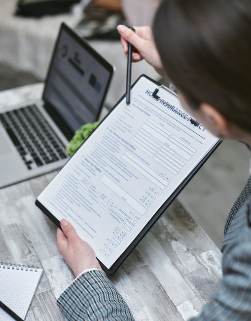 A woman with dark hair styled in an updo, dressed in a beige blazer, stands against a plain white background, holding a clipboard with a visible document titled 'Home Insurance Policy'. She points to the clipboard with a pen in her right hand, indicating a focus on insurance coverage related to house moves. The scene emphasizes the importance of understanding insurance and liability during home relocation or furniture transport, aligning with typical house removal services. The professional appearance and neutral setting support the theme of organised, secure moving logistics.