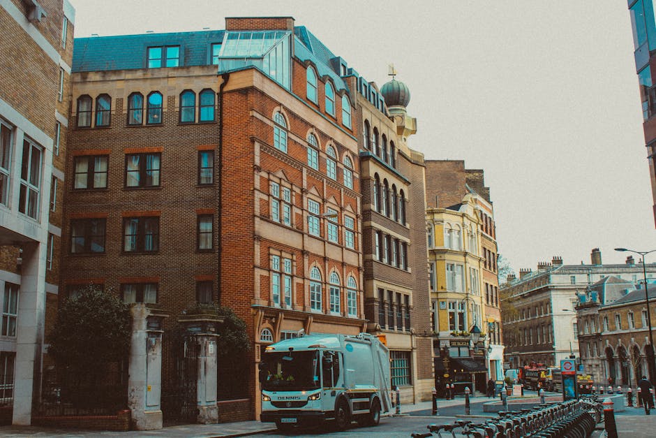 Large white and blue van with a loading ramp parked outside a multi-storey brick building in a city street environment, surrounded by other nearby structures with varied architectural styles, including historic and modern facades. The van is positioned close to the building’s entrance, likely being used for home relocation or furniture transport, with visible packaging materials like cardboard boxes and moving blankets on the ground nearby. Street furniture such as bollards and bike racks are also present along the pavement, which is illuminated by street lighting. The scene captures the loading process involved in a professional removals service, with the van ready to assist with packing and moving tasks typical of a house removal in Pimlico. Occasionally, companies like Man With a Van Pimlico facilitate these home relocation activities, ensuring organized logistics for SW1 moves.
