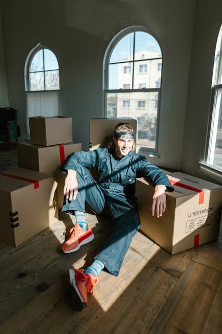 A person dressed in casual clothing and sneakers is sitting on a wooden floor inside a room with large, arched windows that allow natural light to fill the space. The individual is surrounded by several cardboard moving boxes, some sealed with red tape. The boxes vary in size and are arranged around the person, suggesting they are in the process of packing or unpacking during a home relocation. The room has a light-colored wall and a peaceful atmosphere with minimal furnishings visible. This scene illustrates furniture and belongings being prepared for transport, in alignment with house removals and moving logistics. The natural sunlight, the positioning of the boxes, and the person's relaxed posture reflect the stages of packing and loading associated with professional removals services like those provided by Man With a Van Pimlico.