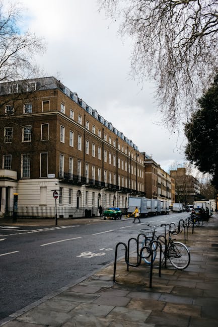 Large white and blue van with a loading ramp parked outside a multi-storey brick building in a city street environment, surrounded by other nearby structures with varied architectural styles, including historic and modern facades. The van is positioned close to the building’s entrance, likely being used for home relocation or furniture transport, with visible packaging materials like cardboard boxes and moving blankets on the ground nearby. Street furniture such as bollards and bike racks are also present along the pavement, which is illuminated by street lighting. The scene captures the loading process involved in a professional removals service, with the van ready to assist with packing and moving tasks typical of a house removal in Pimlico. Occasionally, companies like Man With a Van Pimlico facilitate these home relocation activities, ensuring organized logistics for SW1 moves.