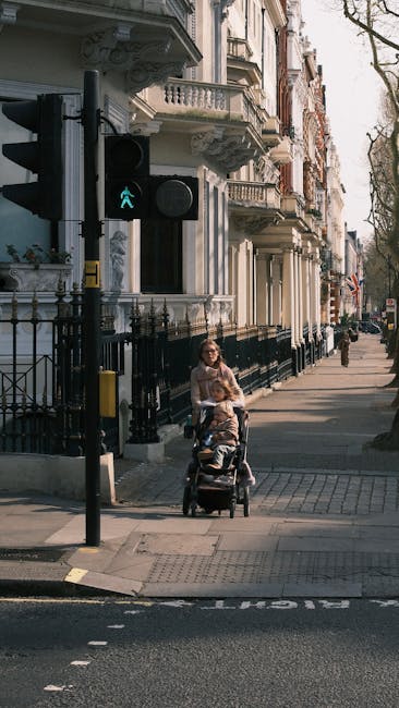 A woman in casual clothing is pushing a stroller with a child along a city sidewalk, passing a pedestrian crossing with a walk signal illuminated in green. The street is lined with historic multi-storey buildings featuring ornate facades, balconies, and decorative ironwork. The buildings are painted in light colors, with some displaying intricate architectural details. A black wrought-iron fence runs along the front of the buildings, and a traffic light with a pedestrian signal is visible on the left side of the image. In the background, another pedestrian is walking further along the sidewalk. The scene is captured during daylight hours, with natural light casting soft shadows across the pavement, creating an urban environment suitable for house removals, local home relocation, and furniture transport services like those offered by Man With a Van Pimlico.