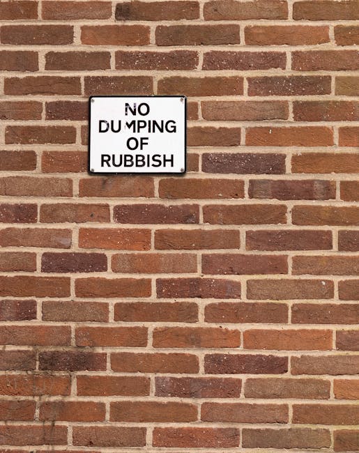 A rectangular white sign with black text mounted on a red brick wall that reads 'NO DUMPING OF RUBBISH'. The wall consists of evenly spaced horizontal rows of bricks with some variation in shades of red and brown, and light grey mortar joints. The sign is positioned centrally in the upper half of the image. The scene is outdoors with natural lighting, highlighting the texture of the brick surface. This image depicts a typical urban boundary restriction that can be relevant in areas near residential or commercial properties involved in house removals or packing and moving services, as provided by Man With a Van Pimlico, especially when managing waste regulations during a home relocation process.