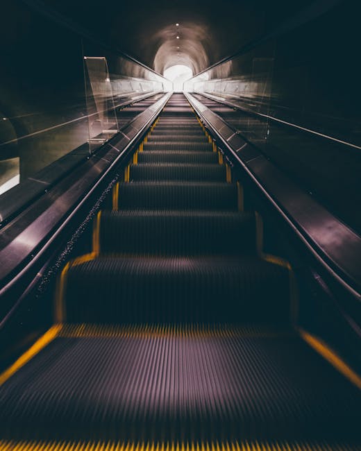 Inside a dimly lit underground pedestrian tunnel with an escalator ascending towards the bright exit. The escalator has black rubber steps with yellow safety edges, and metal handrails on each side. The tunnel’s walls are smooth and dark, with subtle reflections of the lighting. At the top, there is a bright, circular opening revealing daylight, indicating an exit point. The environment suggests a transitional space often used in urban building access or transportation hubs. This scene relates to the logistics of moving and transporting furniture or packed boxes through confined or challenging access areas during home relocation, as managed by Man With a Van Pimlico, emphasizing the importance of careful planning and equipment in house removals and furniture transport.