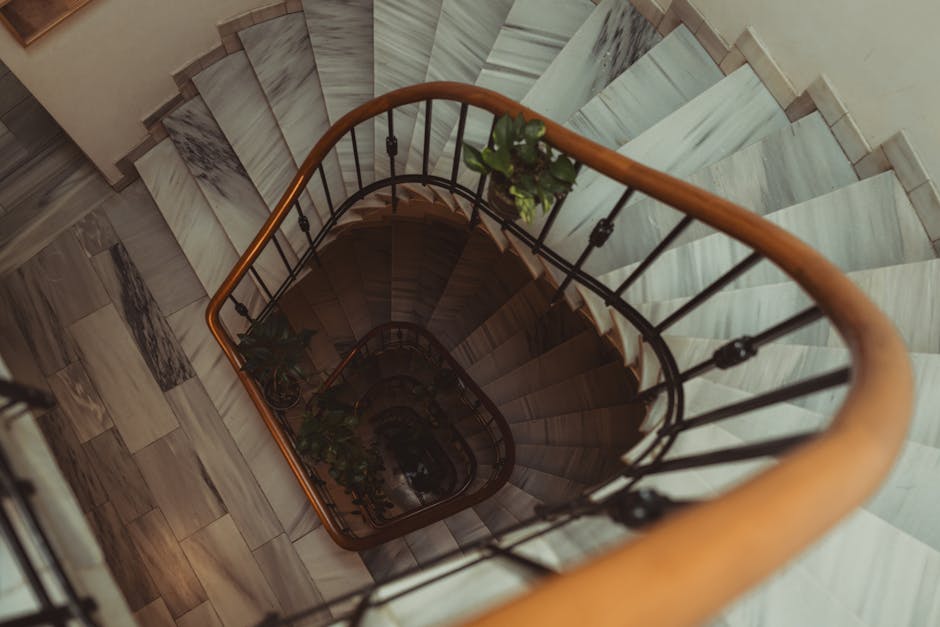 A view from the top looking down a narrow, curved staircase inside a residential property, featuring wooden steps with marble-like surface patterns and a black metal railing with vertical bars and decorative finials, topped by a wooden handrail. The staircase descends to a lower level, partially visible through the open space, with a small green plant positioned on a balcony ledge along the inner curve. The surrounding interior wall is light-colored, and the floor at the top of the stairs is finished with large, rectangular tiles that have a veined marble appearance. This scene captures a typical home interior relevant to house removals and furniture transport, where careful handling and access considerations are essential during a relocation process, as managed by services such as Man With a Van Pimlico.