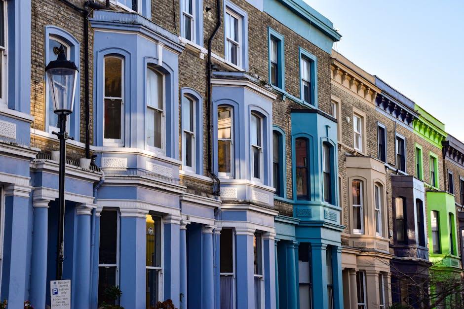 A row of colourful Victorian-style townhouses in Pimlico, London, featuring bay windows with white frames, brick facades, and decorative cornices. The buildings are painted in shades of blue, cream, and green, with some having window sashes and balconies. A black street lamp is visible on the pavement near the flats, with a parking sign beneath it. The scene is captured during daytime with clear skies, and the street appears quiet. These classic terraced houses are part of Pimlico’s residential architecture, often subject to regulations for home relocation and furniture transport, which [COMPANY_NAME] handles with care during house removals in Westminster. The image highlights the urban environment and the building features involved in moving and packing processes, which may require permits according to Westminster Council rules for removals.