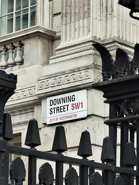 Close-up view of a white street sign mounted on the exterior stone facade of a historic building in Pimlico, London, indicating 'Downing Street SW1' under the jurisdiction of Westminster City Council. The building features detailed architectural elements including stone balustrades and decorative columns, and is partially obscured by a black wrought iron fence with pointed finials and ornamental motifs in the foreground. The scene suggests an urban environment during daylight with natural lighting, where home relocation or furniture transport activities might take place nearby, supported by the presence of a professional removals service such as Man With a Van Pimlico specializing in house removals and moving logistics.