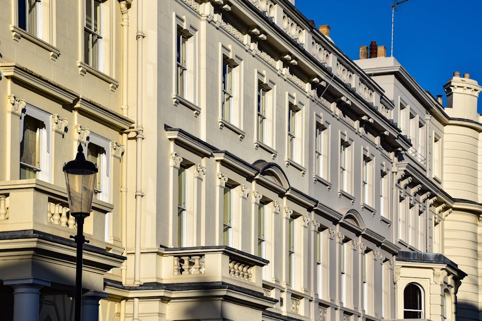A row of colourful Victorian-style townhouses in Pimlico, London, featuring bay windows with white frames, brick facades, and decorative cornices. The buildings are painted in shades of blue, cream, and green, with some having window sashes and balconies. A black street lamp is visible on the pavement near the flats, with a parking sign beneath it. The scene is captured during daytime with clear skies, and the street appears quiet. These classic terraced houses are part of Pimlico’s residential architecture, often subject to regulations for home relocation and furniture transport, which [COMPANY_NAME] handles with care during house removals in Westminster. The image highlights the urban environment and the building features involved in moving and packing processes, which may require permits according to Westminster Council rules for removals.
